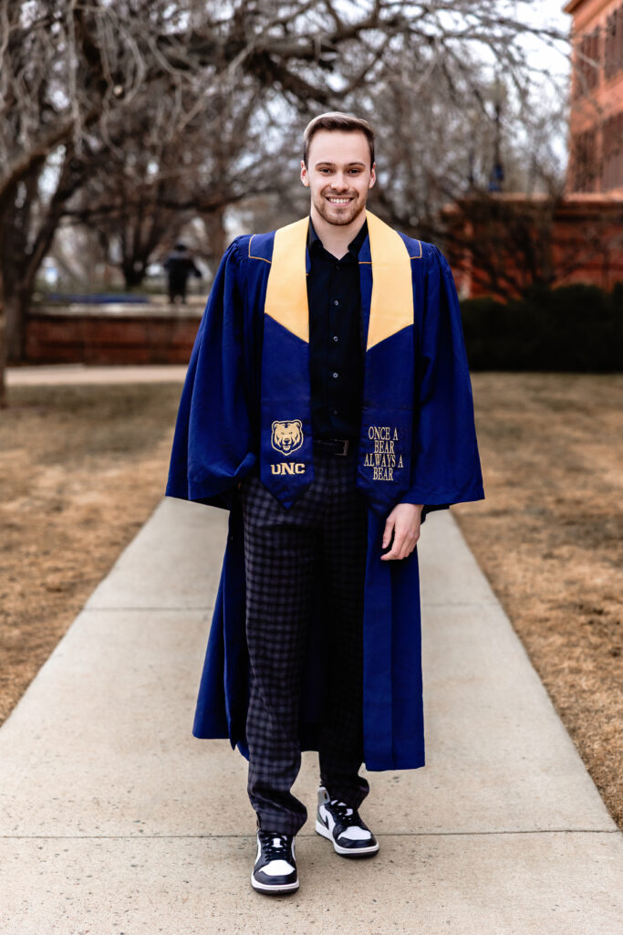 Male college graduate in UNC cap and gown with stole, standing confidently on campus sidewalk in Greeley, Colorado.