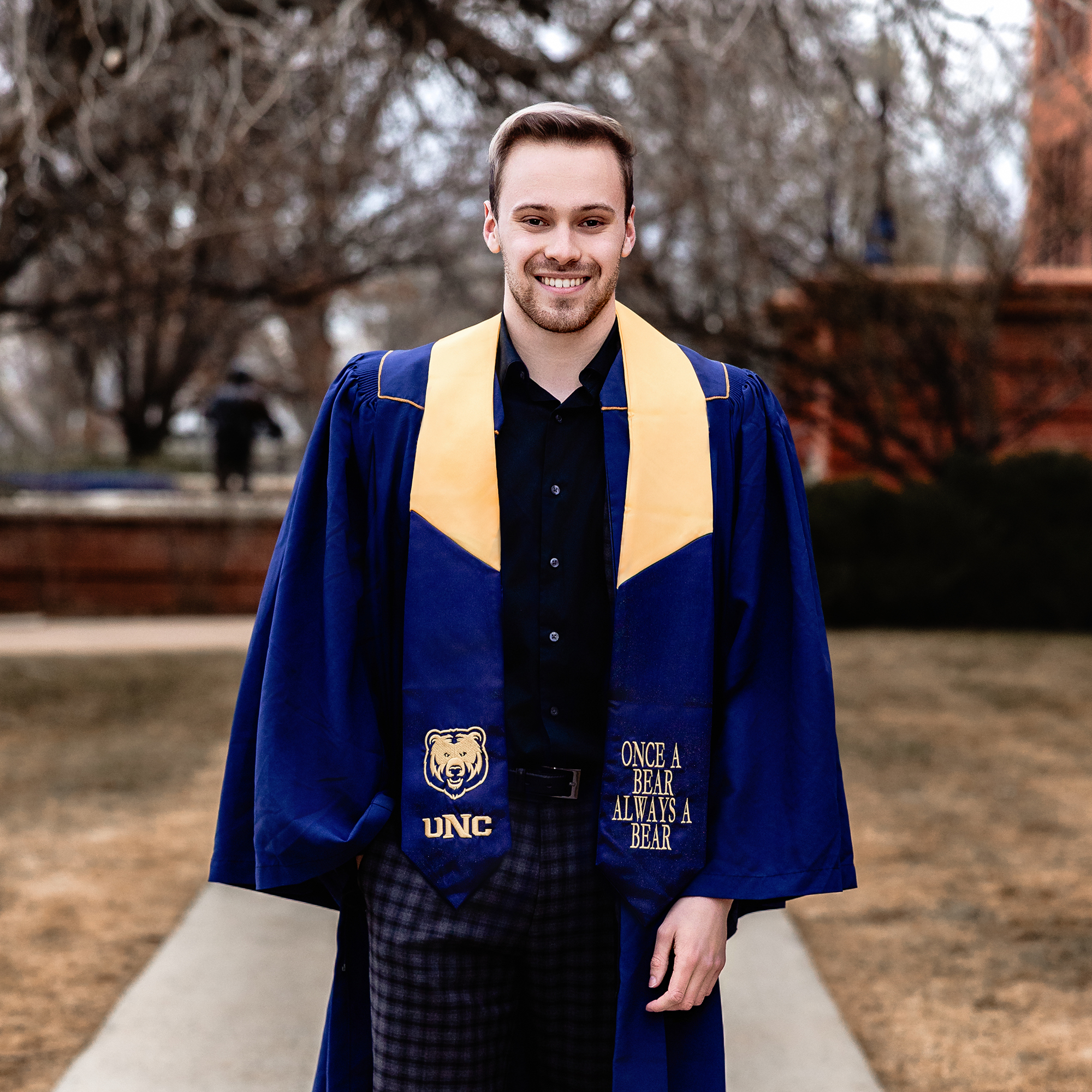 UNC business graduate standing in cap and gown on campus sidewalk in Greeley, CO, wearing a Monfort College stole and smiling confidently.