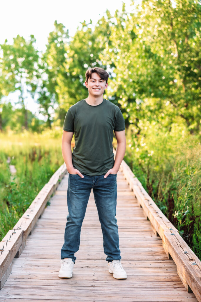 Senior portrait of a teen guy at Signature Bluffs in Greeley, wearing jeans, a green shirt, and white sneakers, smiling confidently with perfect bokeh background