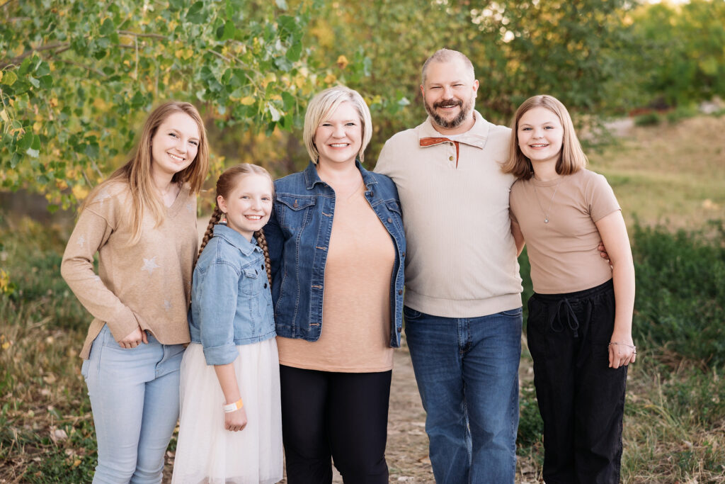 Family of five standing outdoors in coordinated earth-tone outfits for fall portraits, including soft rust, beige, chambray blue, cream, and denim—showcasing a cohesive warm-neutral palette that complements the natural background.