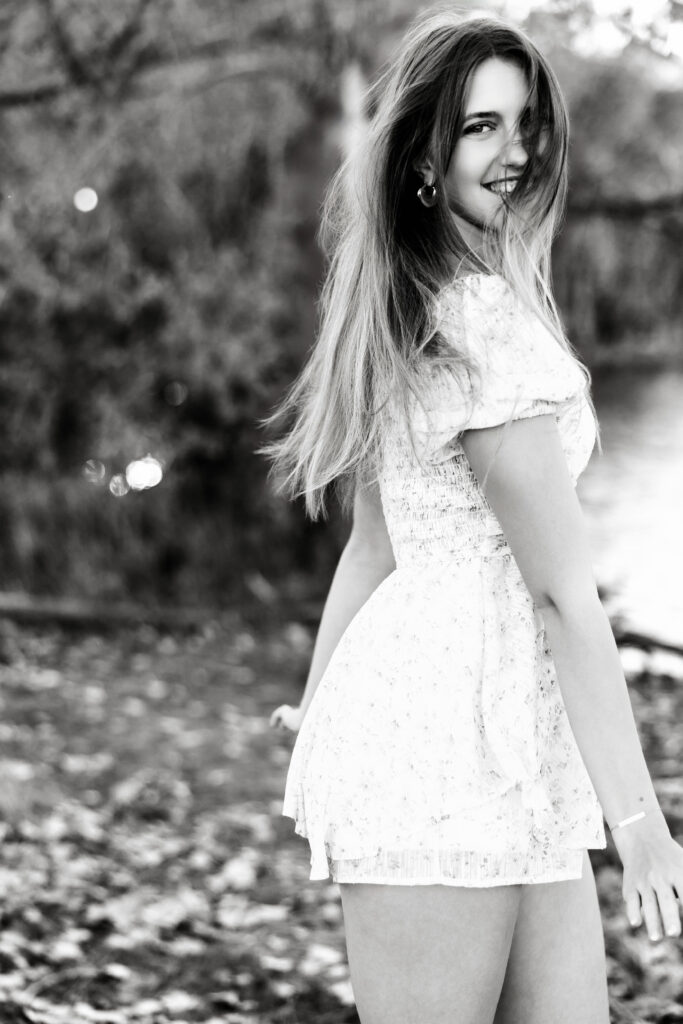 Editorial black and white senior portrait in Greeley CO, high school girl smiling over her shoulder near water with natural wind-swept hair.