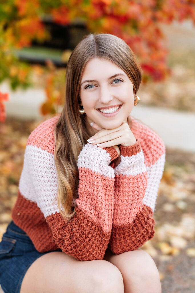 Senior girl sitting outdoors in fall, smiling warmly with her chin resting on her hands, wearing a rust, blush, and cream color-block sweater that highlights her warm skin tone and long light brown hair—framed by colorful autumn leaves in the background.