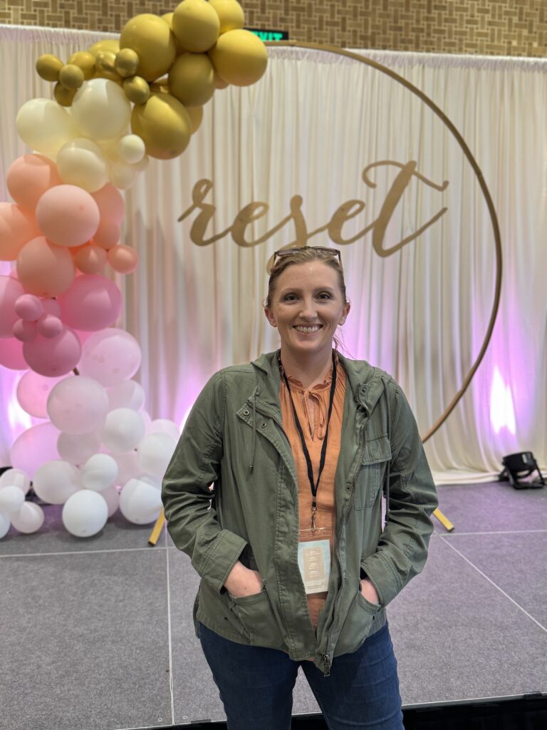 Heather Lilly at the 2025 Reset Conference, wearing a light autumn capsule outfit with a rust blouse, olive green jacket, and dark jeans, standing in front of a balloon arch and “reset” backdrop.