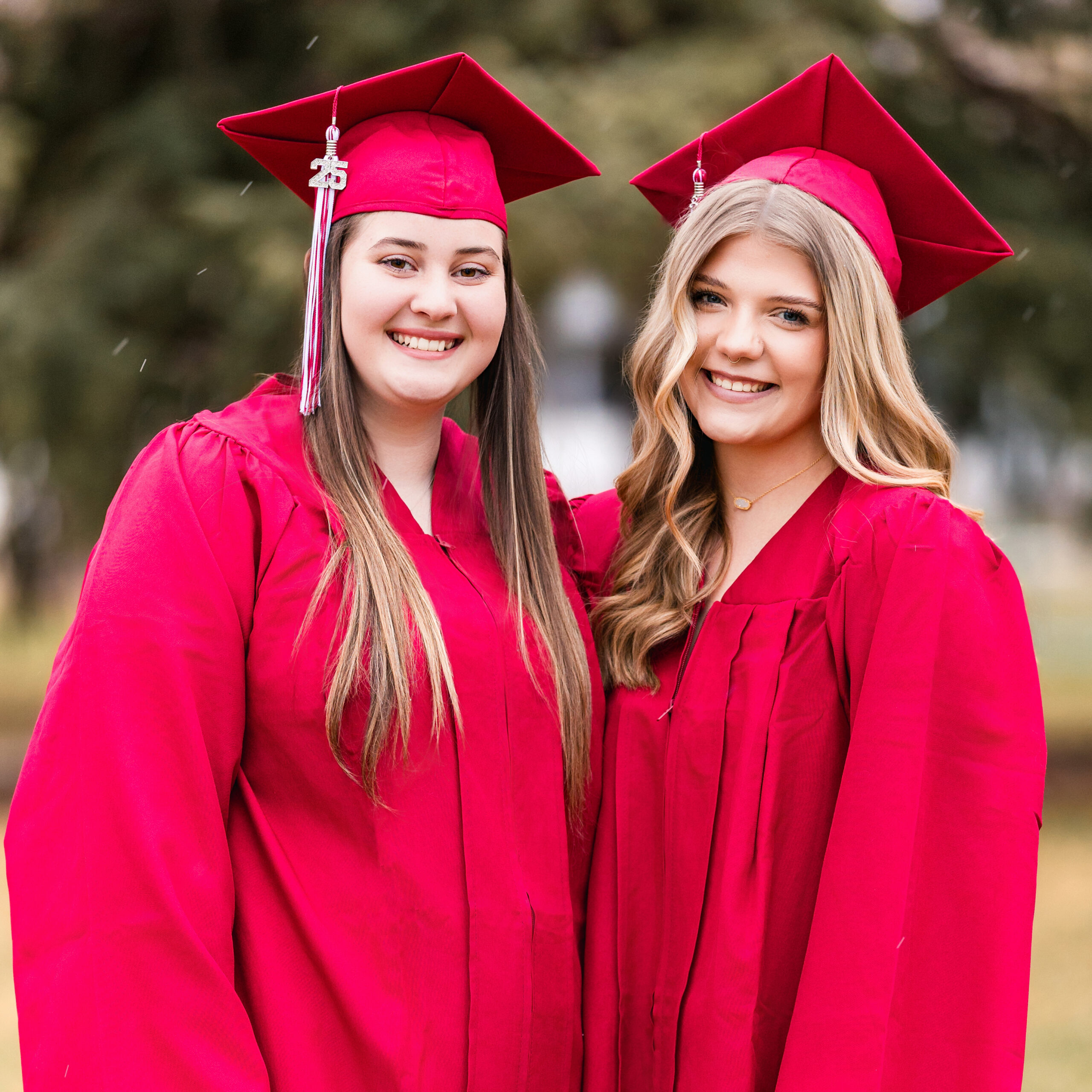 Two high school seniors in red graduation gowns smiling together outdoors during cap and gown photo session in Northern Colorado