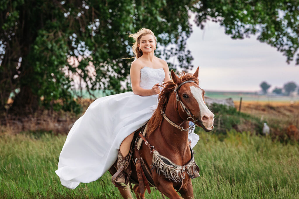 Tween girl in a white bridal gown riding a chestnut horse through tall grass during a milestone equestrian photo session in Eaton, Colorado, captured by Heather Lilly Photography, LLC