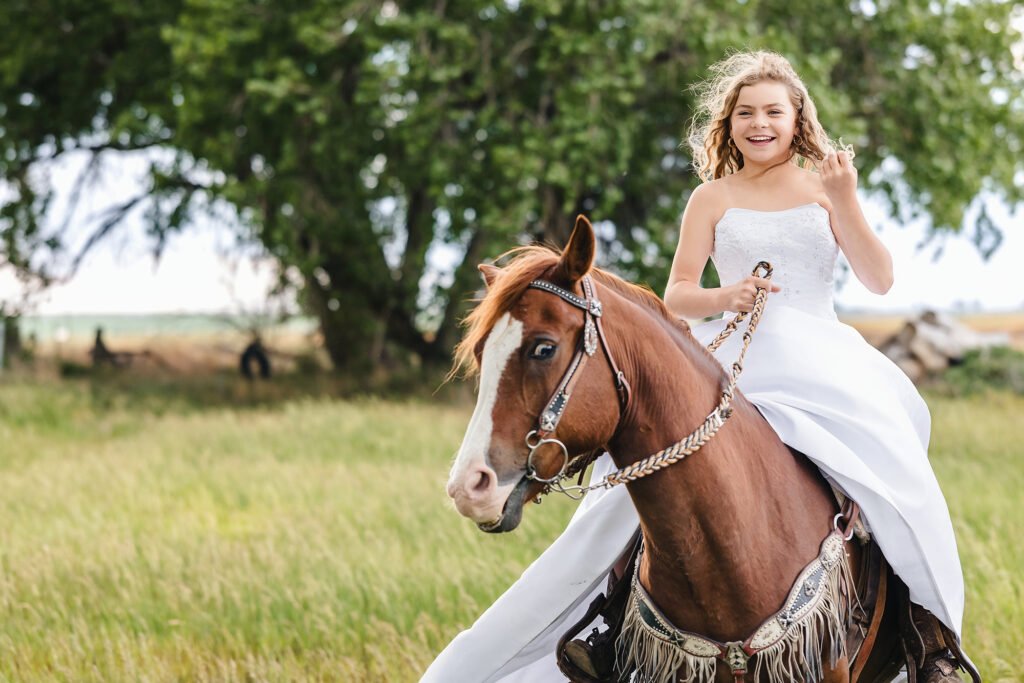 Smiling teen girl riding a chestnut horse in a white wedding gown through a grassy field during a milestone portrait session in Eaton, Colorado, captured by Heather Lilly Photography, LLC
