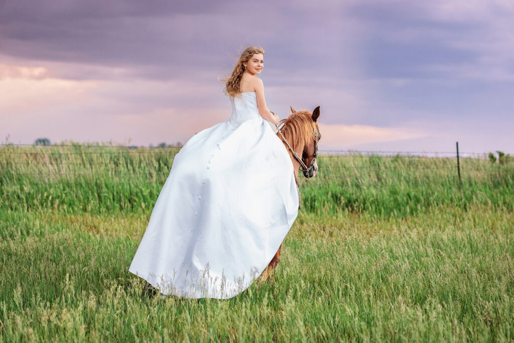 Teen girl in a flowing white dress riding a chestnut horse across a green pasture under dramatic purple sunset skies in Eaton, Colorado with Heather Lilly Photography, LLC