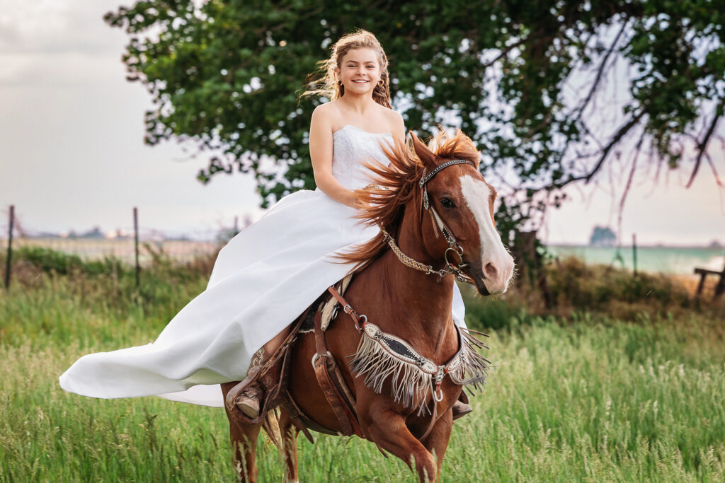 Smiling teen girl riding a chestnut horse in a flowing white dress during an equestrian milestone session in Eaton, Colorado, captured by Heather Lilly Photography, LLC