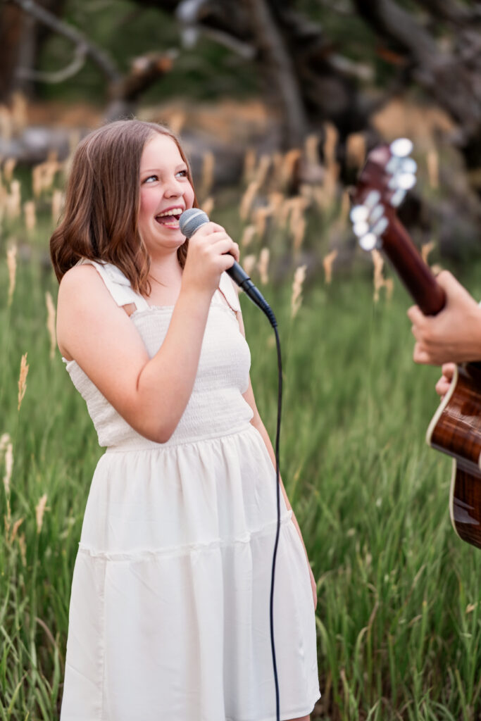 Smiling girl singing into a microphone while her brother plays guitar during a joyful Christian country portrait session in Northern Colorado
