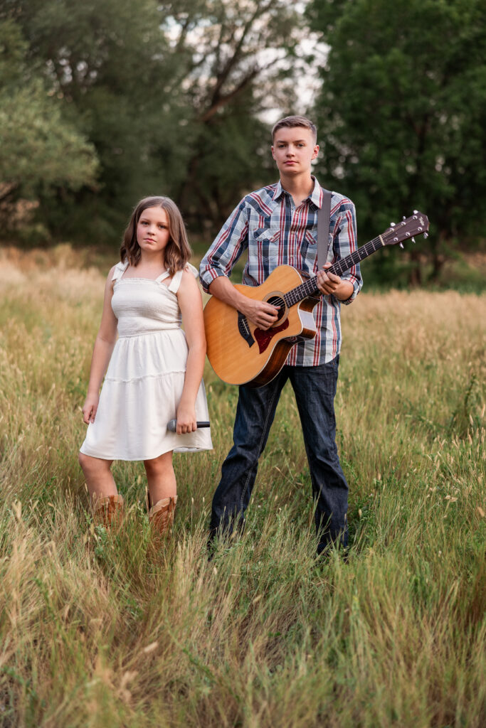 Sibling musicians standing confidently in a golden field with guitar and microphone during an editorial-style country portrait session in Northern Colorado