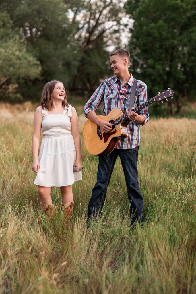 Sibling musicians smiling at each other in a golden field with guitar and microphone during a Northern Colorado country-Christian portrait session