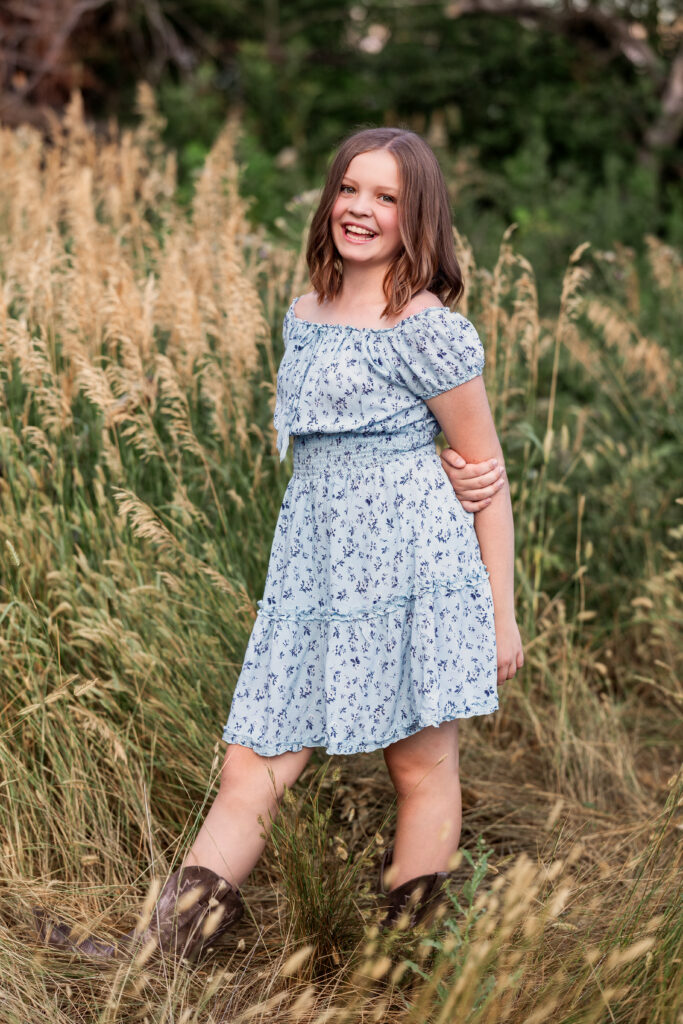 Smiling preteen girl in blue floral dress and cowboy boots standing in tall grass during a country-style portrait session in Northern Colorado