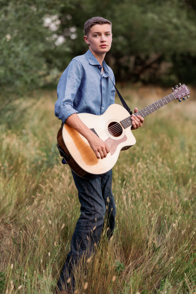Teenage boy holding acoustic guitar in a grassy field during a rustic country portrait session in Northern Colorado