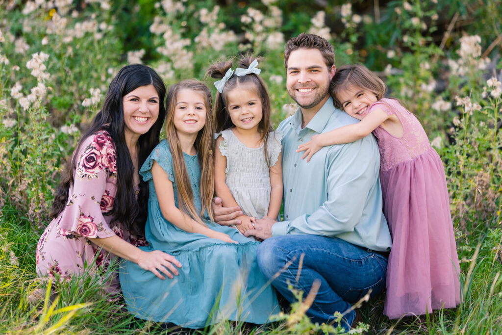 Smiling family of five sitting in a flower-filled field during a summer mini-session in Northern Colorado, wearing coordinated pastel outfits