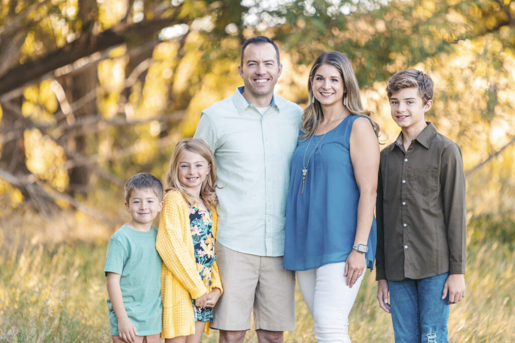 Family of five standing together in golden light during a fall mini-session in Greeley, Colorado, wearing soft earth tones and blues