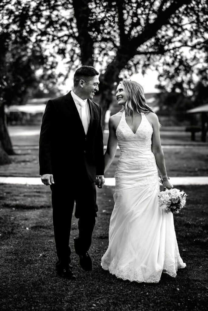 Husband and wife walking hand in hand and smiling during their 15-year anniversary portraits at Lions Park in Cheyenne, Wyoming, captured in black and white by Heather Lilly Photography.