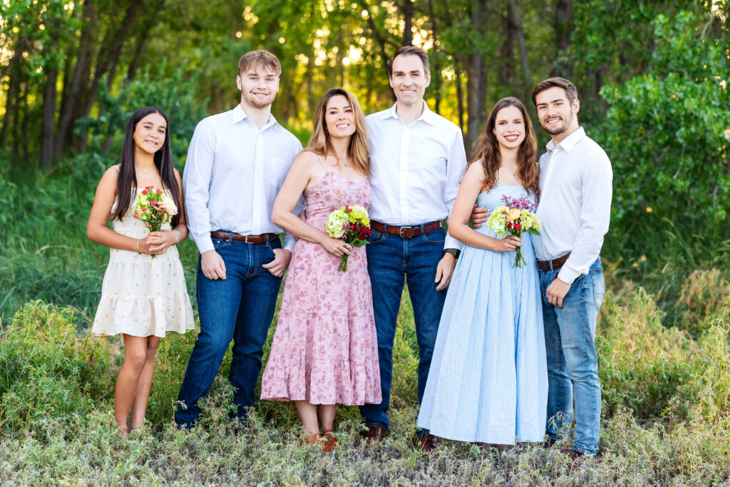 Family of six standing together in tall summer grass at golden hour, dressed in soft neutral and pastel tones, during a milestone photo session in Northern Colorado.