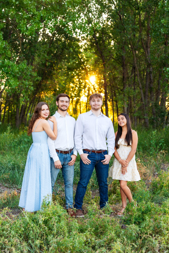 Siblings and newlywed couple standing together in tall summer grass, smiling during golden hour at a family photo session in Northern Colorado.