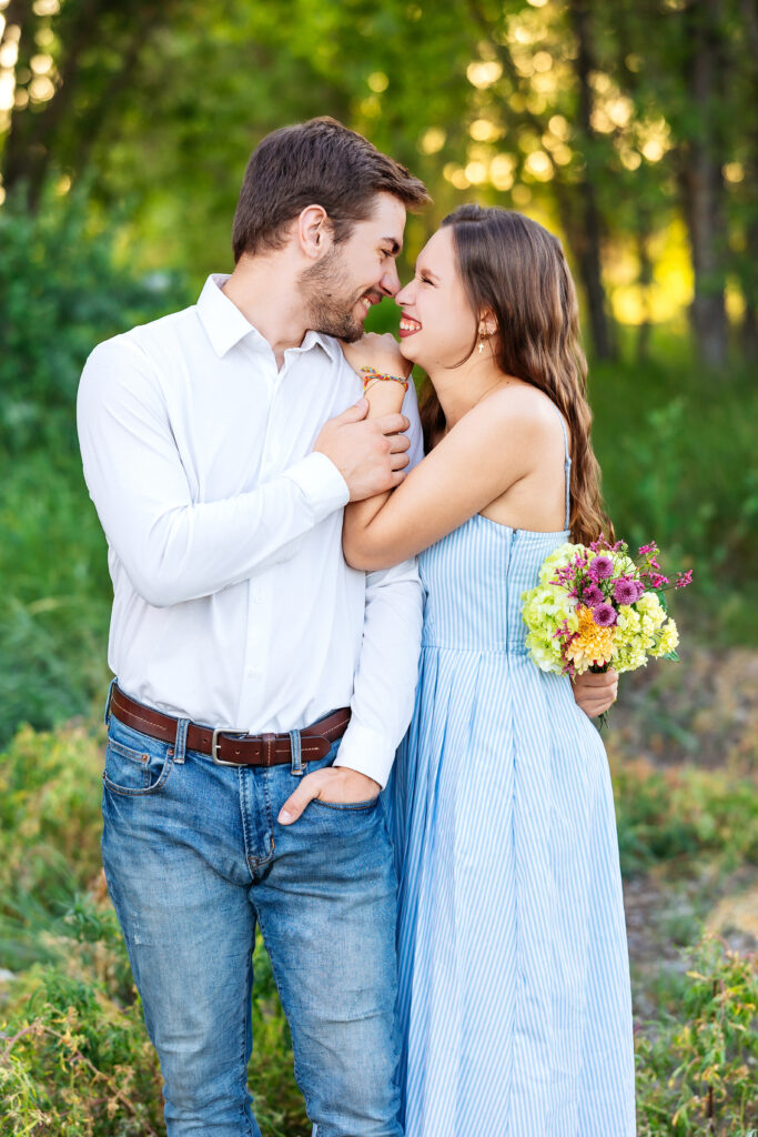 Young couple smiling with foreheads touching, holding a bouquet of summer flowers during a golden hour family session in Northern Colorado.