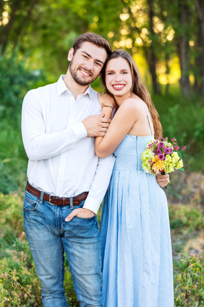 Young couple smiling and embracing in a sunlit forest during a Northern Colorado summer family session, holding a bouquet of wildflowers.