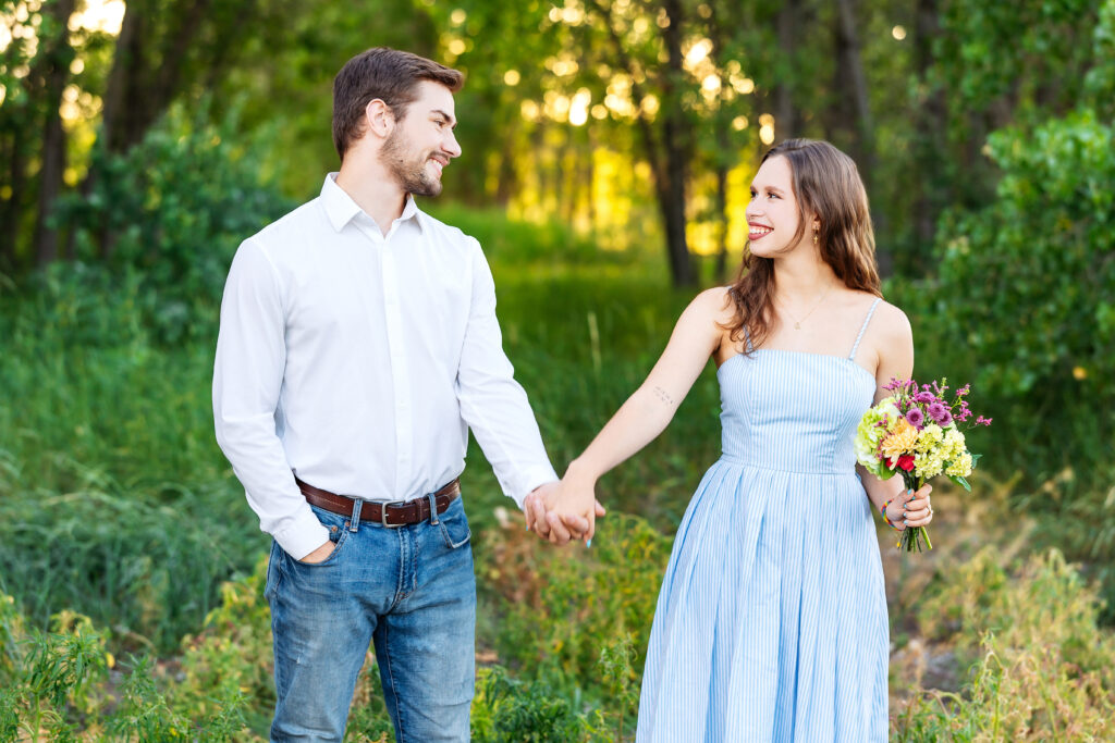 Young couple holding hands in tall grass, smiling at each other during golden hour in a Northern Colorado family photo session.
