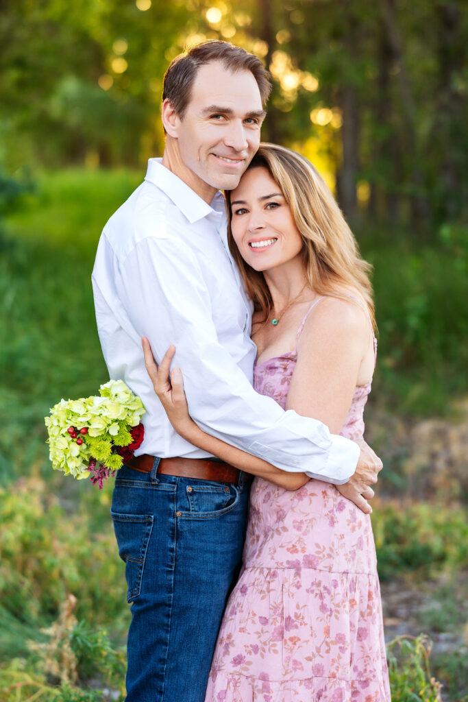 Husband and wife embracing closely and smiling softly during a golden hour family session in Northern Colorado, surrounded by trees and natural light.