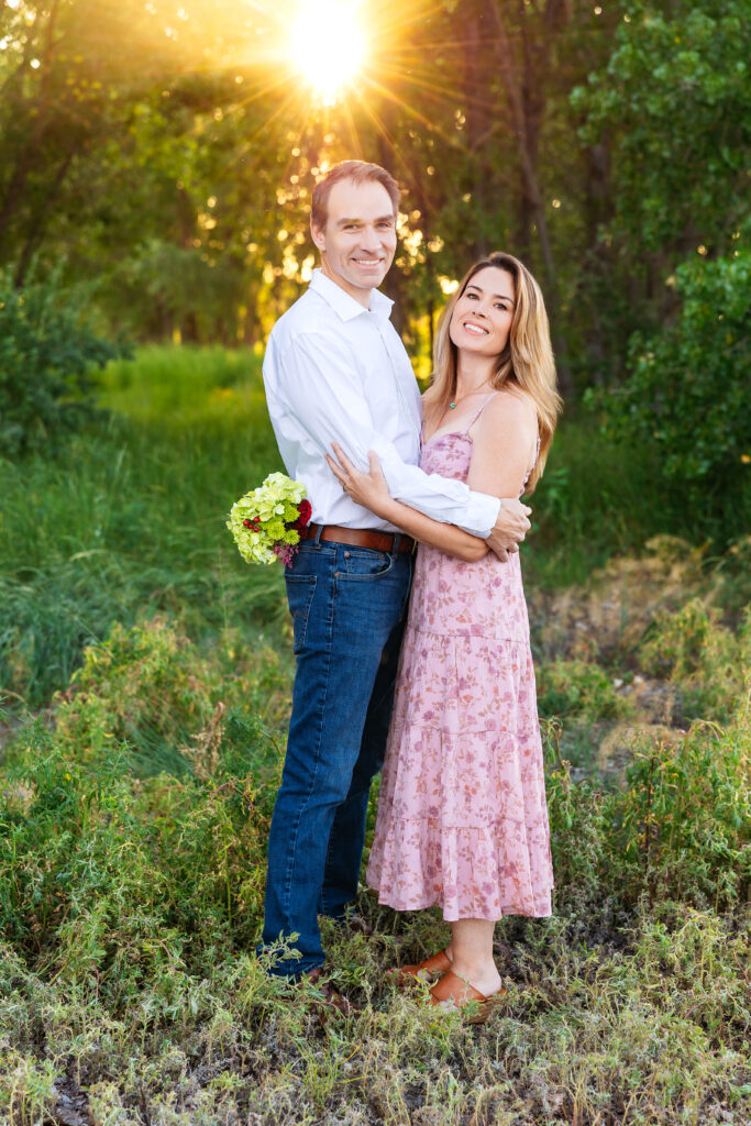 Husband and wife embracing in golden hour light, with a big sun flare, standing in tall summer grass during a milestone family session in Northern Colorado.
