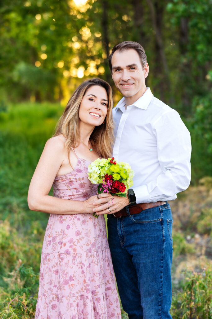Couple smiling softly during family portraits, holding a floral bouquet with green and burgundy tones in a natural Northern Colorado setting.