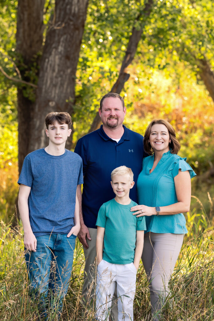 Family standing in tall grass wearing coordinating blue and green outfits for their portrait session near Greeley, Colorado with Heather Lilly Photography.