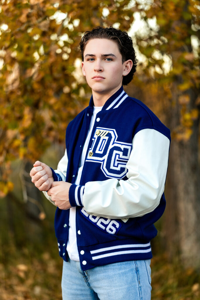 High school senior portrait of a student wearing a navy letterman jacket in golden autumn light, photographed in Northern Colorado.